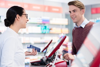 Side-view of a cheerful pharmacist holding two packs of prescribed medicine while giving advices, and useful information to a young male customer in a modern drugstore.
